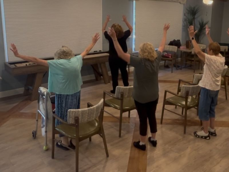 A group of three seniors behind teacher standing behind chairs in a community room, participating in a adaptive dance class with both arms raised overhead. A walker is visible near one participant, and shuffleboard-style tables and large windows with cellular shades are in the background.