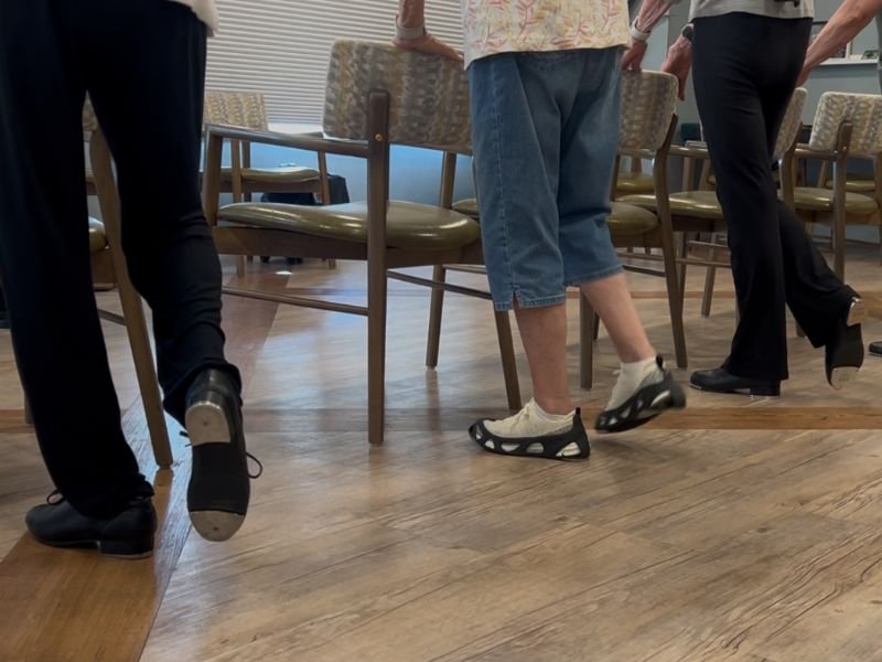 Close-up view of three seniors' legs and feet during a chair-assisted standing tap class, each standing behind a chair with one foot lifted or extended in a leg exercise. The wood-look flooring and rows of chairs are visible in the background.