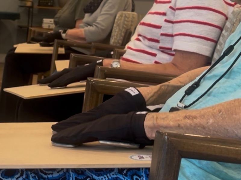 Row of seated people at small desks, each wearing a black fingerless glove with a metal tap plate on the palm, using it to tap-tap rhythms on the desk surface in front of them.